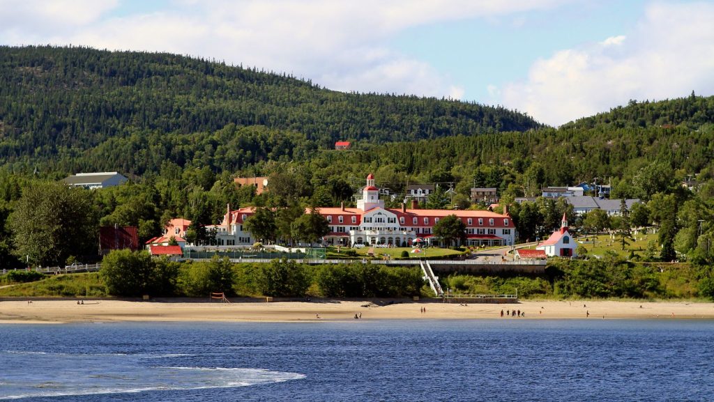 Vue aérienne du pittoresque village de Tadoussac niché entre une verdure luxuriante et un littoral pittoresque, avec l'emblématique baie de Tadoussac visible en arrière-plan. Des bâtiments pittoresques et des monuments historiques sont dispersés dans tout le village, créant une atmosphère charmante et sereine.